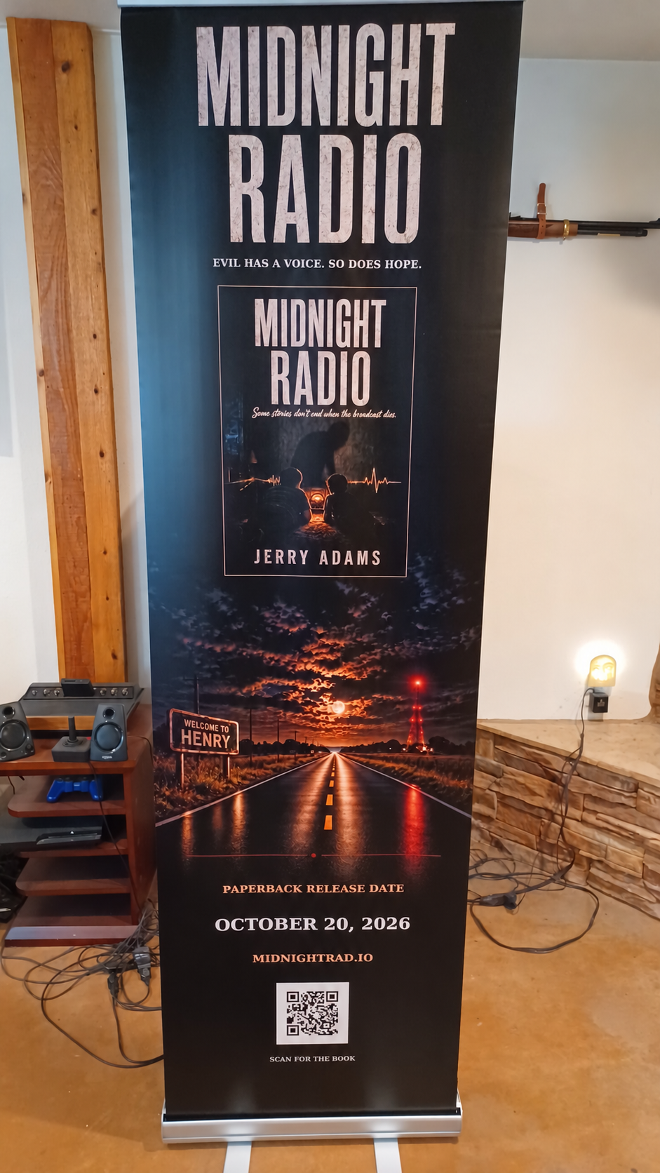 Jerry Adams’s seven-foot Midnight Radio book-signing banner at home, beside his gun cabinet and Henry rifle.